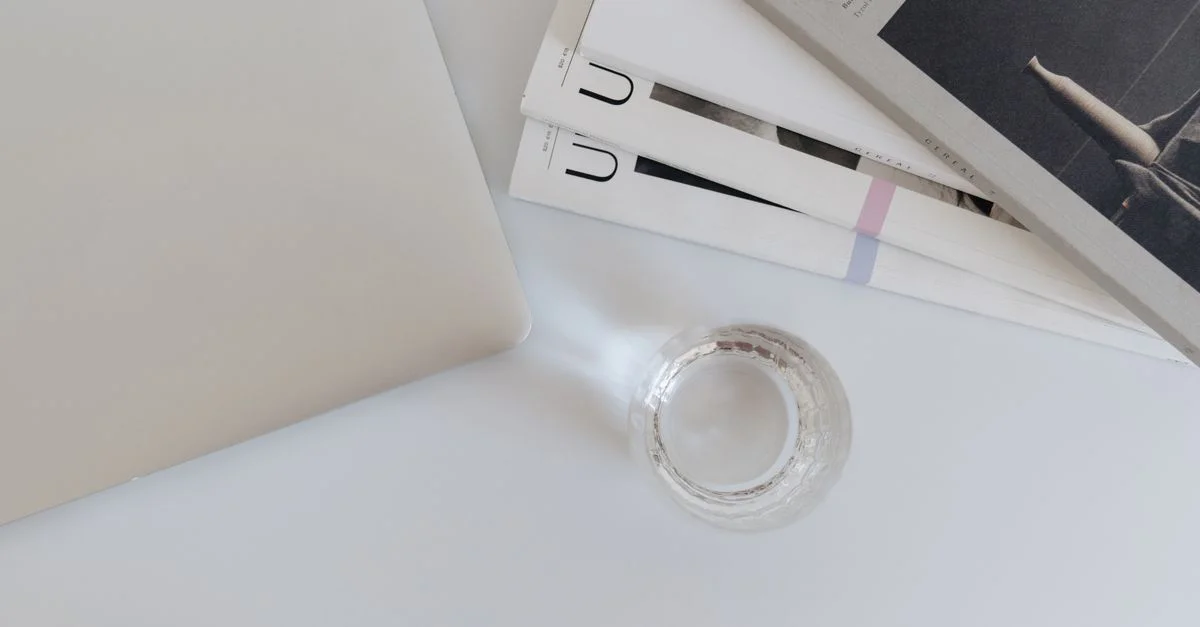 Un espace de travail propre et minimaliste avec un ordinateur portable, des magazines et un verre sur un bureau blanc.