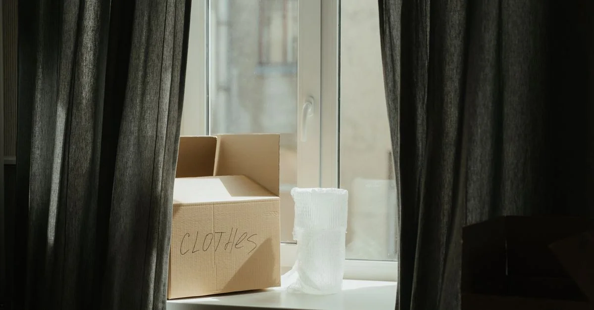 Boîte en carton avec des vêtements à côté d'une fenêtre, idéale pour des thèmes de déménagement ou de relocalisation.